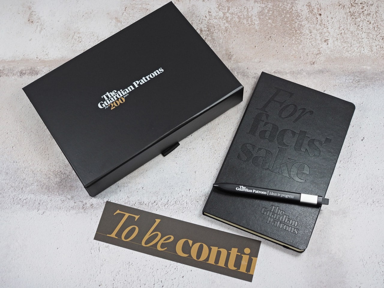 A branded moleskine pen sits on top of a debossed moleskine notebook beside a custom bookmark as part of the patron gift sets.